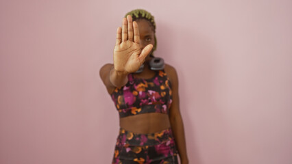 Woman with braids performs stop gesture against a pink wall wearing vibrant workout attire,...