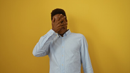Fototapeta premium Young man with hand on face over isolated yellow background wearing blue shirt looking through fingers