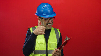 Man with helmet and vest holds clipboard against red background showing determination and confidence in construction industry