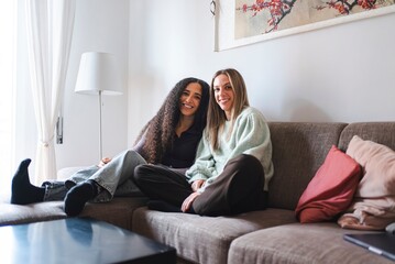 Two young women sit closely on a couch, smiling warmly at the camera, appearing happy and relaxed in a cozy indoor setting with natural light coming from a window.