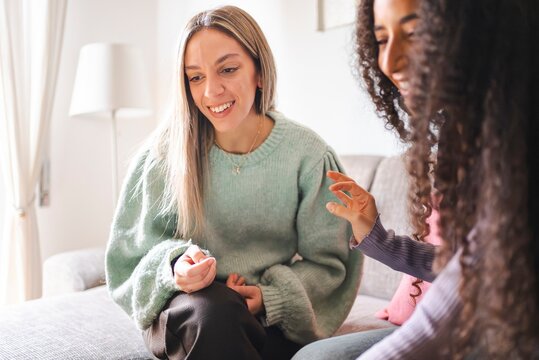 A woman in a mint green sweater smiles while sitting on a couch, listening to her curly-haired friend, as they share a moment of warmth and connection in a cozy home setting.