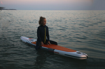 An elderly woman rides paddleboarding in the ocean. Senior female is waiting for a wave on a surfboard. Active old age. Old girl is sitting on a board in the sea at sunset, dressed in a swimming suit