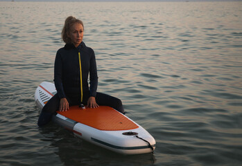 An elderly woman rides paddleboarding in the ocean. Senior female is waiting for a wave on a surfboard. Active old age. Old girl is sitting on a board in the sea at sunset, dressed in a swimming suit