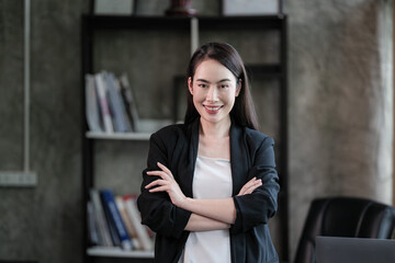 A confident asian businesswoman stands with arms crossed in a modern office, wearing a blazer and smiling professionally at the camera.