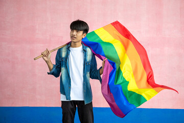 Asian young man holding colorful rainbow flag on pink color background. Male with a gay pride flag,...