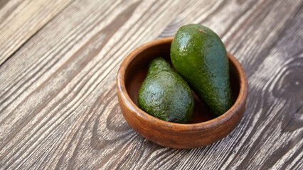 Two ripe green avocados placed in a round wooden bowl on a textured rustic wooden surface, captured in natural light with visible grain patterns