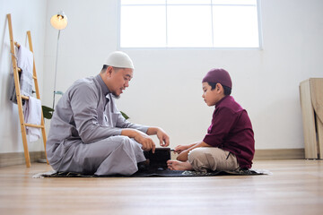 Side View Of Asian Muslim Male Teaching Son To Reading Quran While Sitting On Prayer Mat
