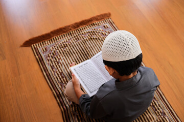 Rear View High Angle Of Muslim Kid Reciting Holy Qur'an On Prayer Mat