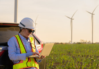 Female engineer wearing hard hat and safety vest using laptop while leaning on car in wind farm. Caucasian woman working outdoors in green field with wind turbines. Renewable energy and technology