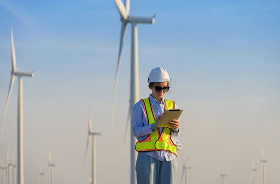 Female wind farm engineer in safety vest working on tablet with wind turbines in background. Caucasian woman in white hard hat inspection of renewable energy installation