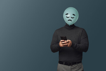 Man with smartphone hiding behind balloon with sad face on dark grey background. Space for text