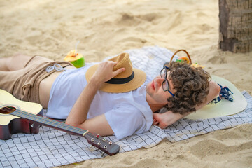 Carefree tourist enjoys a peaceful afternoon nap on a sandy beach with a guitar by his side. Surrounded by nature, he reflects the ultimate slow travel experience and pure relaxation...