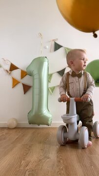 Cute baby boy celebrates one year birthday. Toddler riding a toy balance bike in sunny room among balloons and sage green inflatable number one. Child in beautiful green suit.