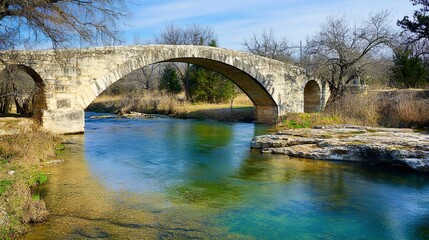 Fototapeta premium A picturesque stone bridge arching over a clear, flowing stream, its weathered surface complemented by the natural beauty of the water below. 