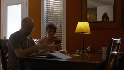 A middle-aged man and his senior mother sit together at a dining table in a warmly lit home, sorting through papers and bills. Son counts receipts, while mother is onlooking. 24 fps.