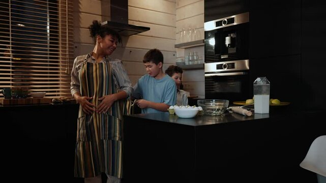 Two sons stand and tie an apron on their mother who is standing in front of them, then one of them cuts butter with a knife, in the kitchen