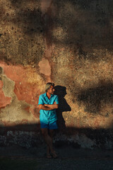 Man with blonde hair in blue shirt and swimming shorts in late afternoon sunlight leaning against old pink stucco wall.