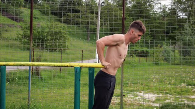 Young sportsman doing push ups on parallel bars at nature. Strong sporty man exercising at sports ground. Male shirtless athlete training hard outdoor. Concept of active sportive lifestyle. Slow mo