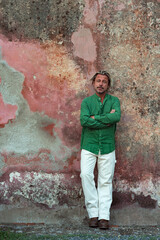 Obraz premium Tanned man with blonde hair in green shirt and white pants leaning against old pink stucco wall.