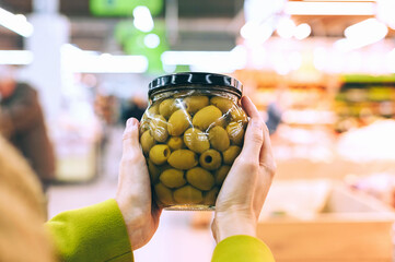Hands holding glass jar of green olives in supermarket aisle.