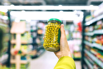 Hand holding jar of green peas in supermarket aisle.