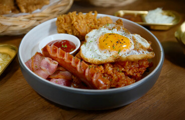 Hearty breakfast bowl with fried rice, sunny-side up egg, sausage, bacon, and fried chicken, served with ketchup in a white bowl on wooden table.