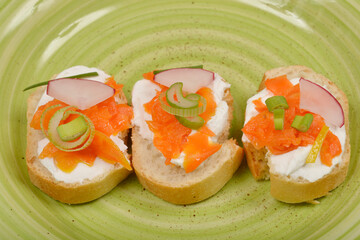 Three small open-faced sandwiches feature a creamy spread topped with vibrant orange carrot prepared as vegan salmon, thin radish slices, and fresh green chives on a textured light green plate