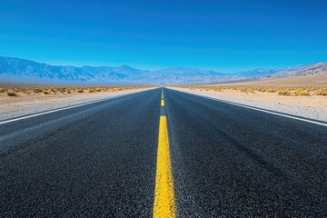 Naklejka premium Empty highway stretches into a vast, desert landscape under a vibrant blue sky. Mountains loom in the distance
