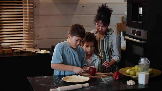 Mother standing at the table and letting her two sons cut an apple, in the kitchen