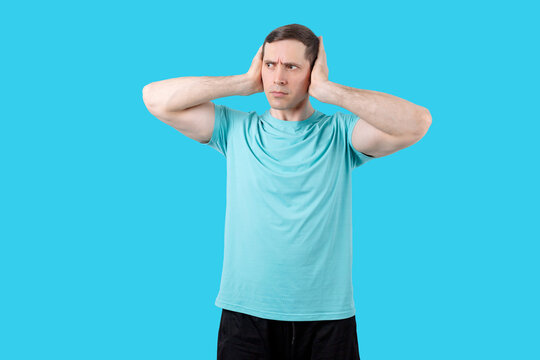 Young man in blue t-shirt covers ears with hands discontentedly and gets angry on blue background. Isolated studio portrait. People and emotions concept Young man exercising with dumbbells