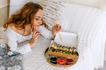 A young beautiful woman eating a sushi set of rolls in a takeaway box while sitting on a bed in a...