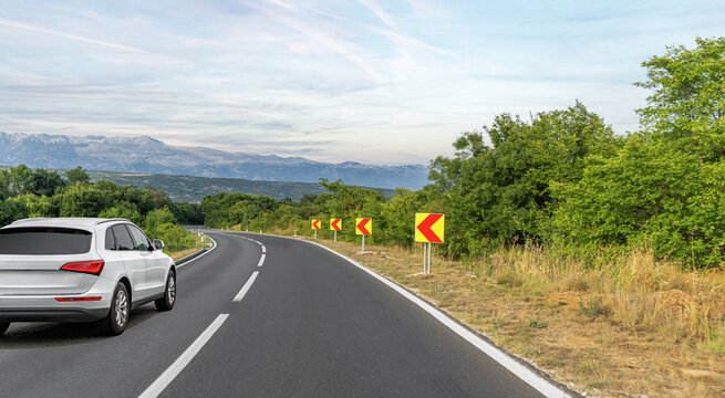 A white SUV drives along a curving countryside road with sharp turn warning signs, surrounded by green trees and mountain views under a cloudy sky.