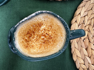 Crafted cup of salted caramel coffee sits on a textured green surface with a woven placemat adding warmth. Creme brulee delicate crust is visible