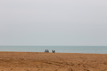 The image shows a beach scene with a vast expanse of sand in the foreground and the ocean in the background. There are four individuals riding bicycles on the sand near the water's edge. Bushehr, Iran
