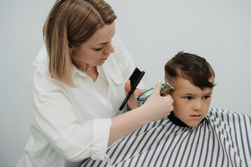 A female hairdresser cuts a little boy's hair with a clipper. A professional hairdresser and a child client in a modern barbershop