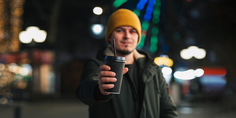 A Caucasian man stands outside on a winter evening with a paper cup of coffee in his hands.