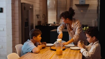 A multiracial adult woman cuts and serves pie at a wooden kitchen table while two school-aged boys drink juice and wait with plates in a modern home.