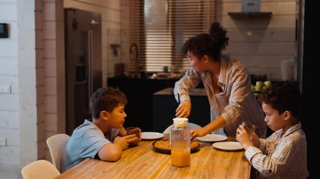A multiracial adult woman cuts and serves pie at a wooden kitchen table while two school-aged boys drink juice and wait with plates in a modern home.