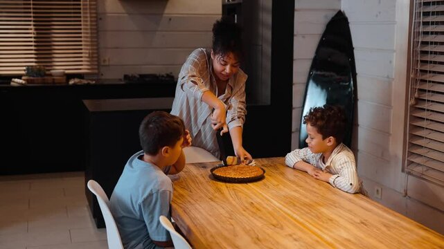 An adult Black woman is standing near a dining table and cutting a pie while her two school-age sons are sitting at the table across from each other, waiting for their piece of pie.