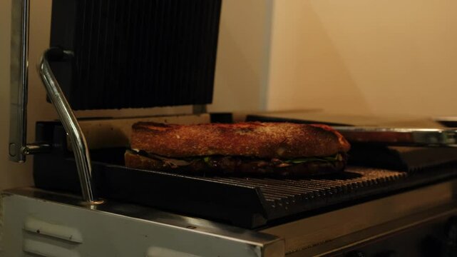 The chef slowly opens the lid of the electric grill and takes a toasted, delicious meat sandwich on a baguette with his hands. Warm grilled sandwich preparation, beautiful food, lunch idea.