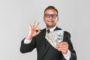 Cheerful businessman in a suit holds american dollars in his hands and shows an ok gesture with his...