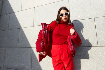 High fashion photo of a young brunette woman in red glossy down jacket, and red pants. Urban street...