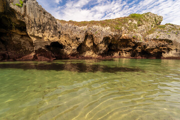 Natural rock arch rises from the shallow waters of a golden sandy beach at Playa de Cuevas del Mar in Asturias, Spain.