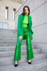 High fashion photo of a young brunette woman in green coat, poloneck and leather pants.