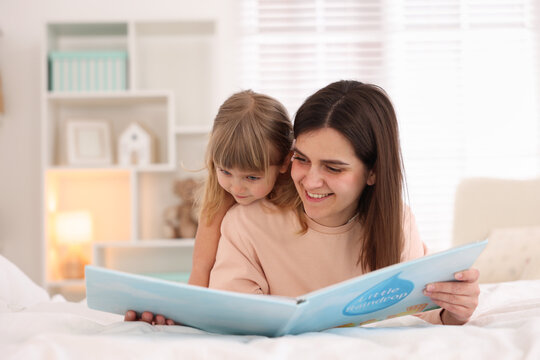 Bedtime. Cute little daughter reading book with her mother on bed at home - Powered by Adobe