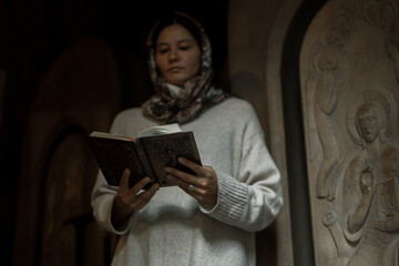 Woman with Asian-European features in headscarf and white dress praying and lighting candles in an...