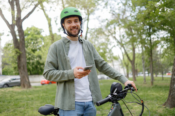 Smiling man in green helmet enjoys cycling in park while using smartphone on sunny day