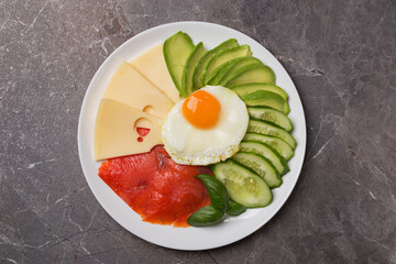 Keto breakfast plate with scrambled eggs, salmon, cheese, cucumbers and avocado on dark table. Healthy eating plate.