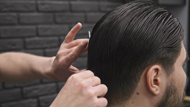 A young man with brown hair has his hair cut with thinning scissors at the back of his head at a barbershop.
