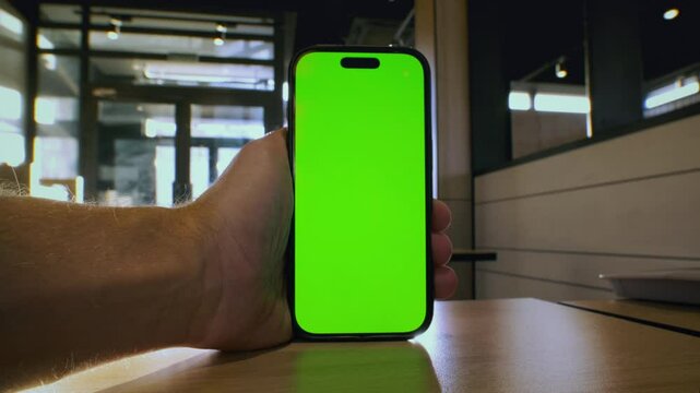 POV: Man holds a smart phone in his hands with a green screen for chroma key in cafe, restaurant. Male using gadget with app for delivery. Food court. Guy zoom in map on the phone display